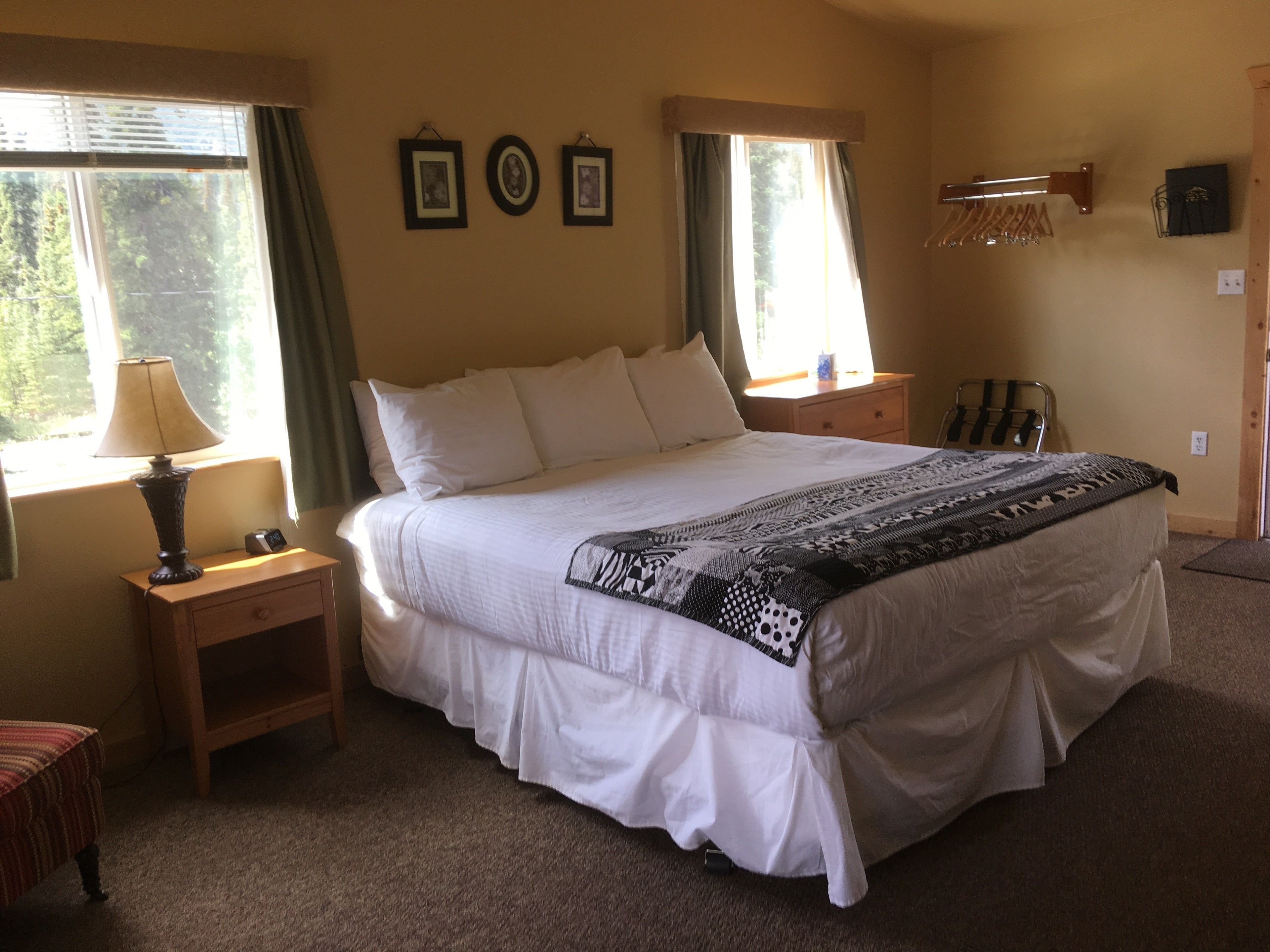 A neatly made bed with a patterned throw, a bedside lamp, and a nightstand. There are two windows with curtains, a wall-mounted coat rack, and a small shelf with a lamp.
