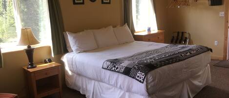 A neatly made bed with a patterned throw, a bedside lamp, and a nightstand. There are two windows with curtains, a wall-mounted coat rack, and a small shelf with a lamp.