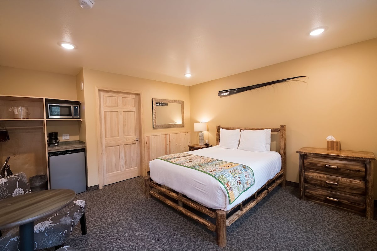 A bedroom with a wooden bed, a wooden nightstand, a bedside table, a microwave, a refrigerator, and a wooden dresser.