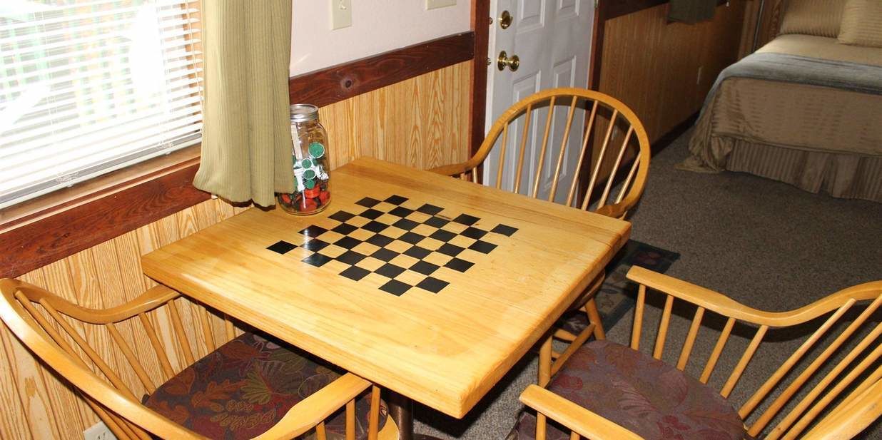 A wooden table with a checkered game board, surrounded by wooden chairs with cushioned seats.