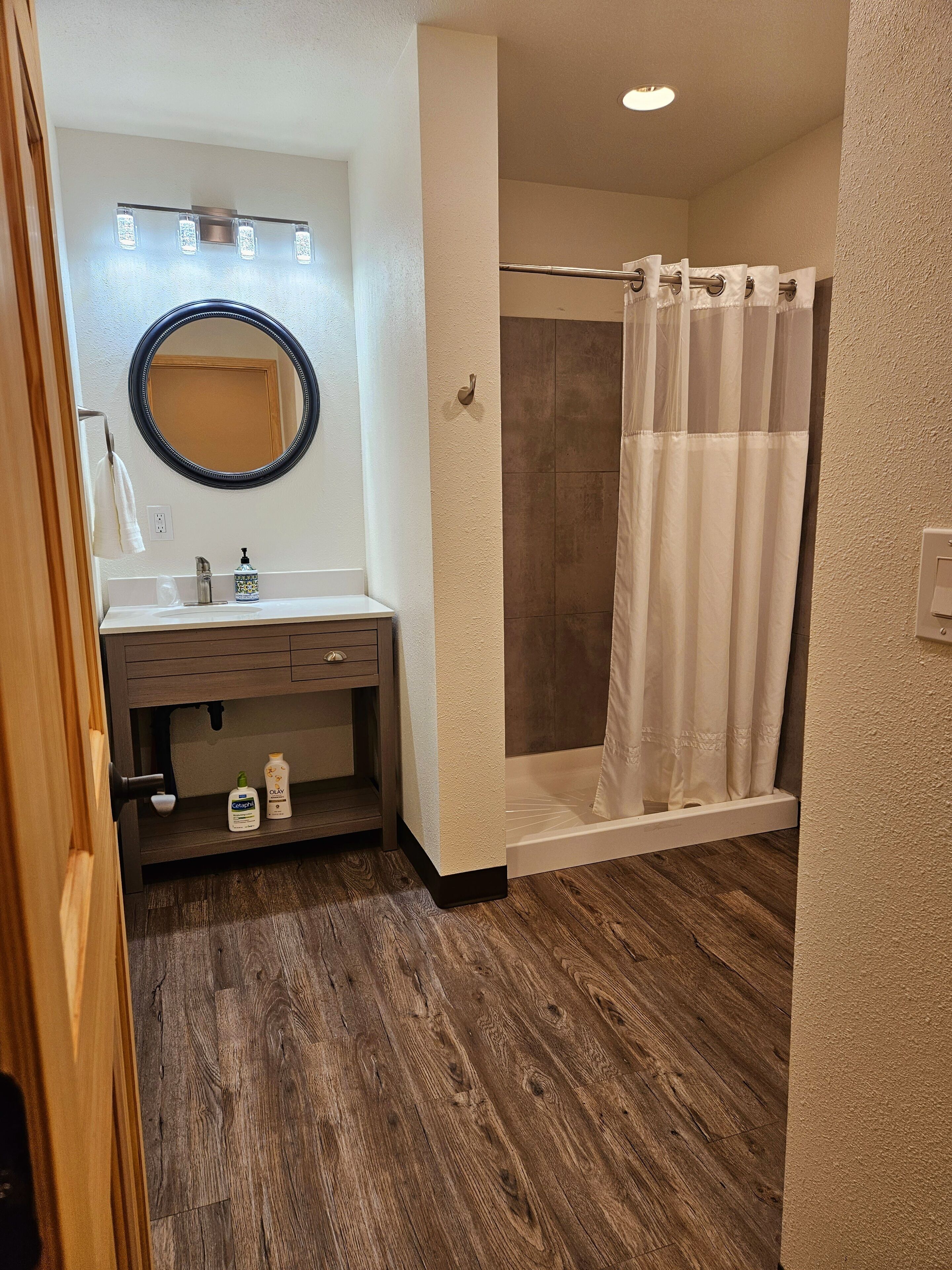 A bathroom with a wooden floor, a sink with a mirror, a shower with a curtain, and a towel rack.