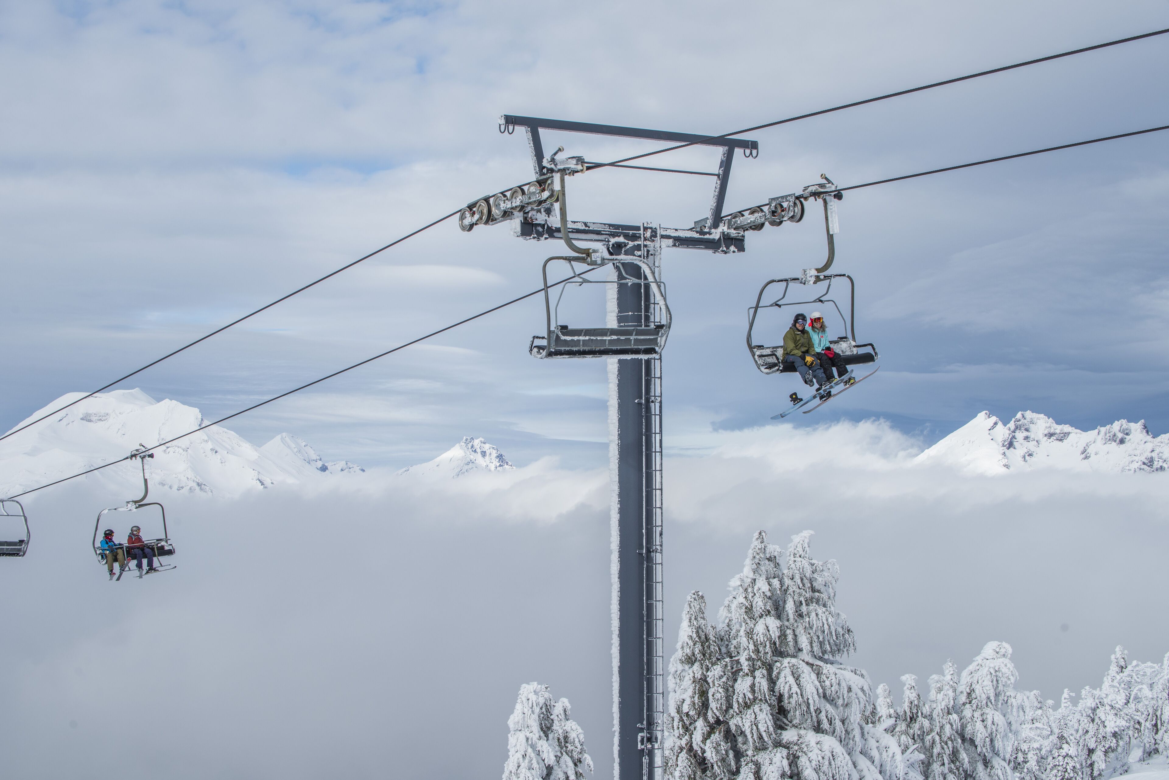 En skilift med stole, der transporterer skiløbere foran sneklædte bjerge og træer.