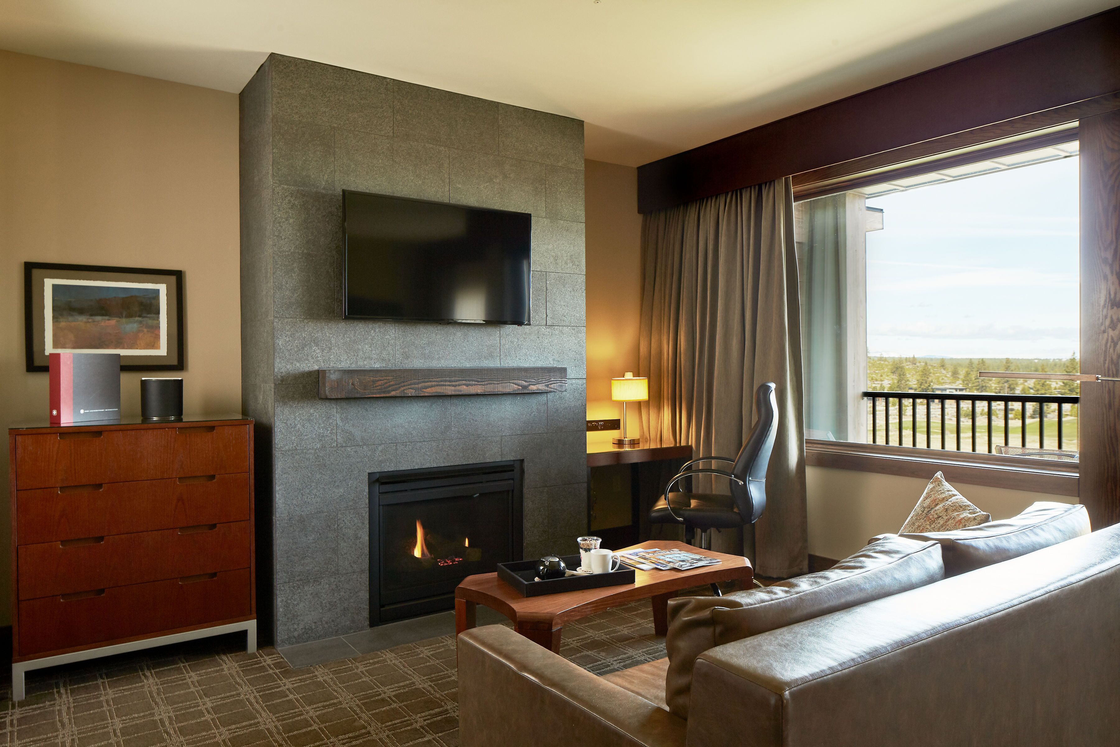 A modern hotel room with a fireplace, a flat-screen TV mounted on the wall, a sofa, a wooden coffee table, and a view of the outdoors through a large window.