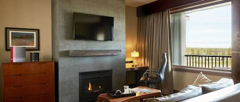 A modern hotel room with a fireplace, a flat-screen TV mounted on the wall, a sofa, a wooden coffee table, and a view of the outdoors through a large window.
