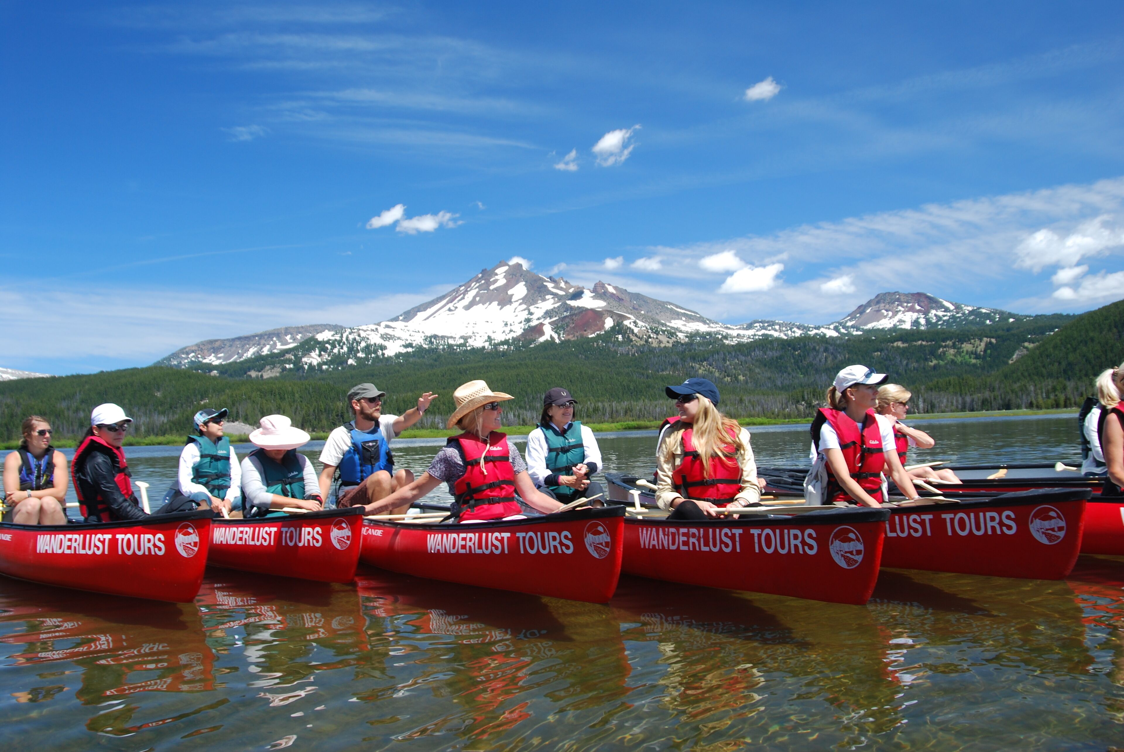 A group of people in red canoes labeled "Wanderlust Tours" on a calm lake with a mountain in the background.