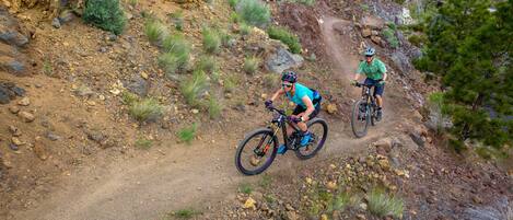 Dois ciclistas de montanha numa trilha de terra, rodeados por terreno rochoso e vegetação rala.