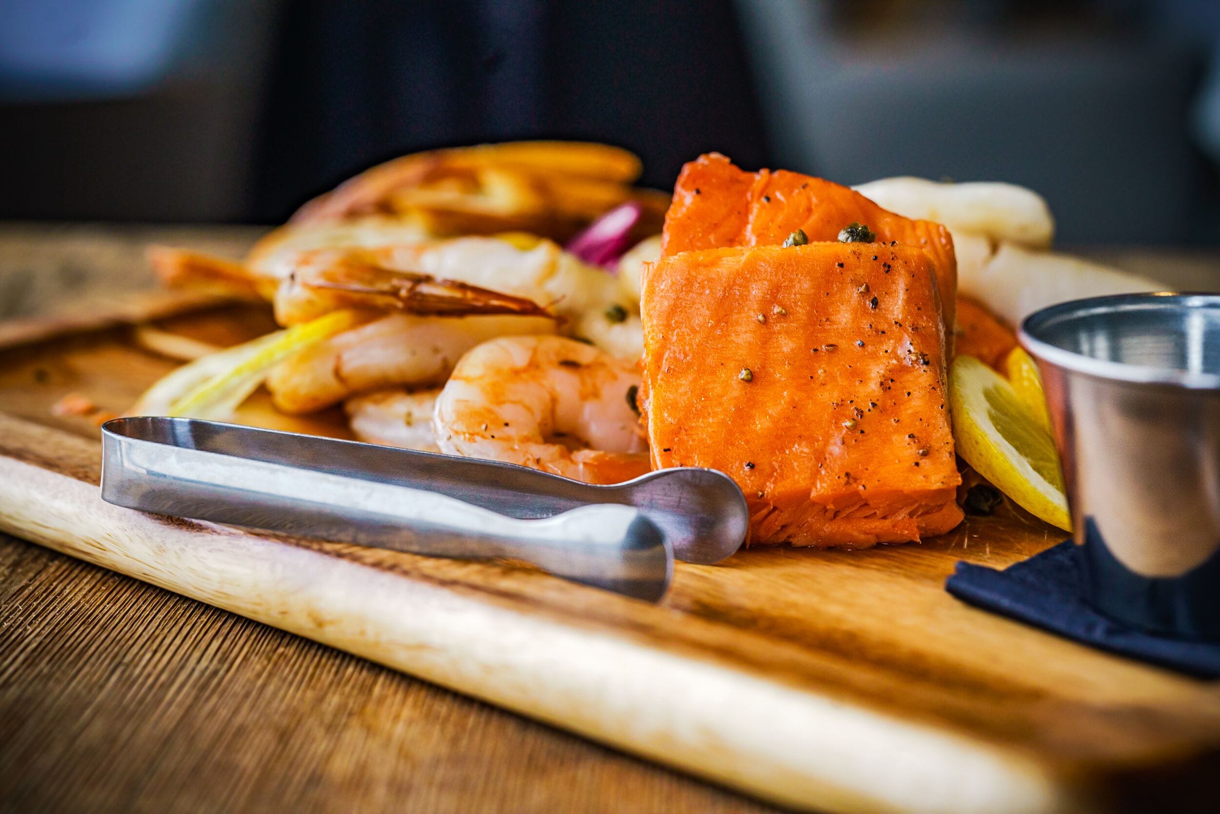 A wooden cutting board with a piece of salmon, shrimp, and lemon slices.