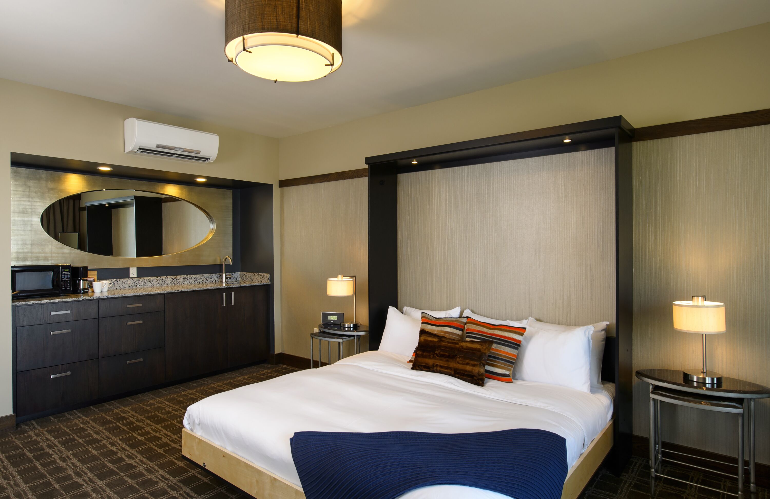 A hotel room with a large bed, a round mirror, a built-in air conditioning unit, a small bedside table with a lamp, and a dark-colored wardrobe.