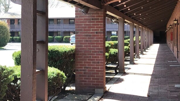 A covered walkway with brick pillars and a tiled floor, leading to a building with a balcony. - Lincoln Lodge (Urbana)