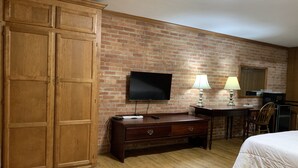 A hotel room with a brick wall, wooden wardrobe, a flat-screen TV, a desk with a lamp, a bed, and a microwave. - Lincoln Lodge (Urbana)