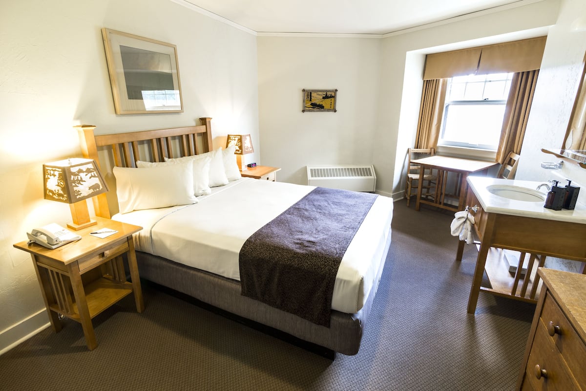 Une chambre d’hôtel comprenant un lit, une table de chevet avec une lampe, un bureau avec une chaise, une fenêtre avec des rideaux et une salle de bain avec un lavabo et un miroir.