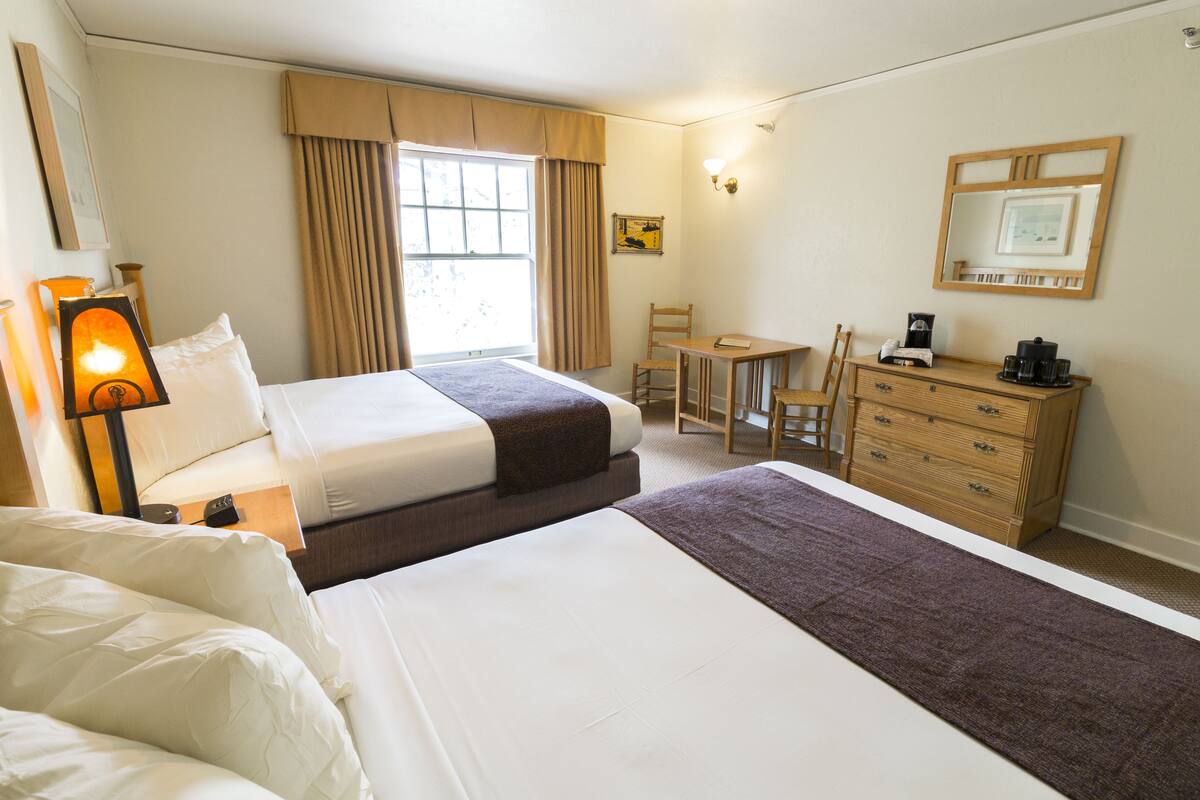 Une chambre d’hôtel avec deux lits, un bureau, une chaise, une commode et une fenêtre avec des rideaux.