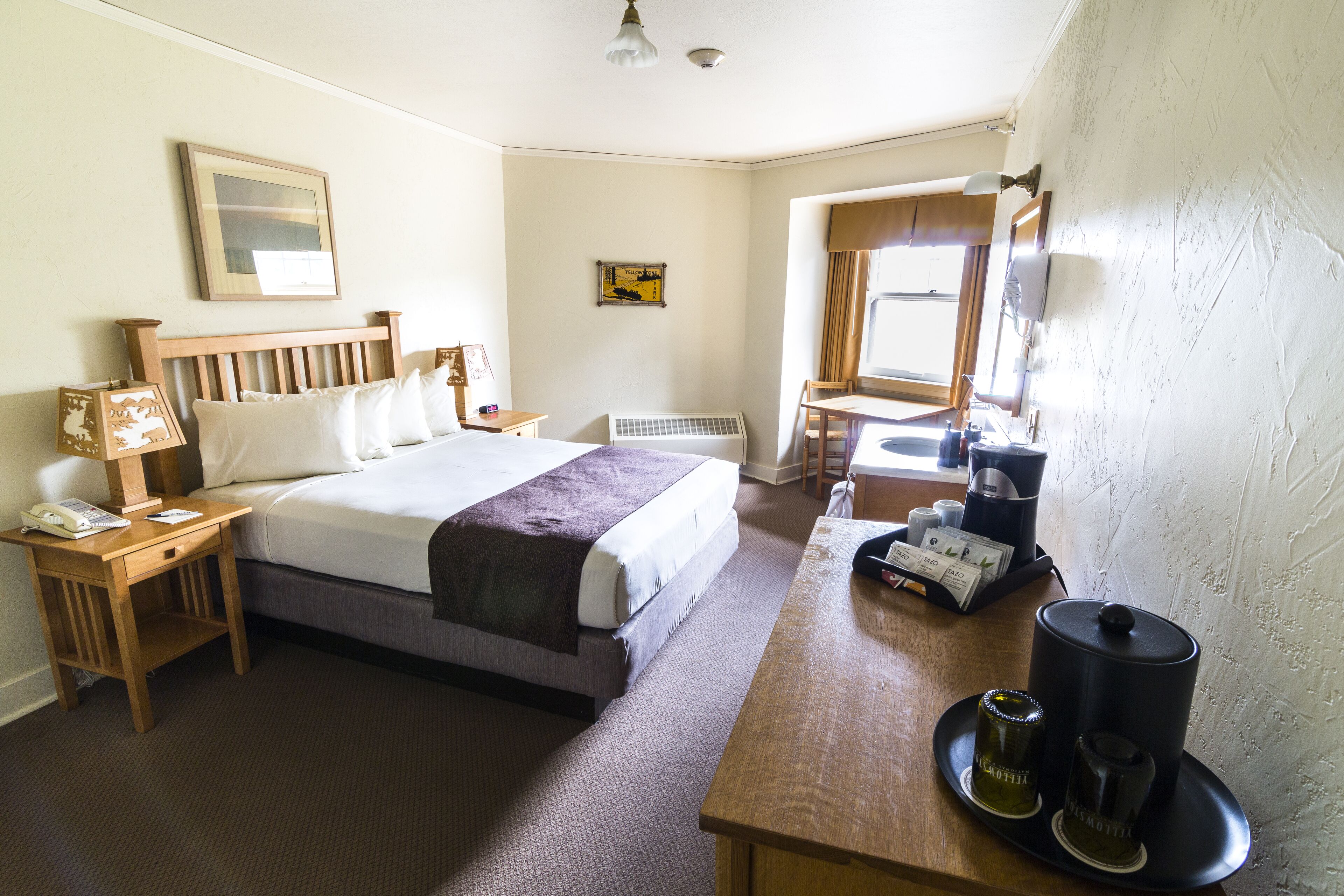 A hotel room with a bed, bedside table, lamp, and a small table with a coffee maker and cups.