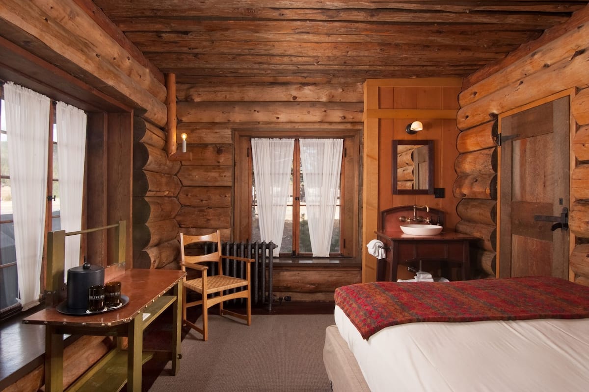 Une chambre de style rustique, avec des murs en rondins, un bureau en bois, une chaise, un lit et une fenêtre avec des rideaux.