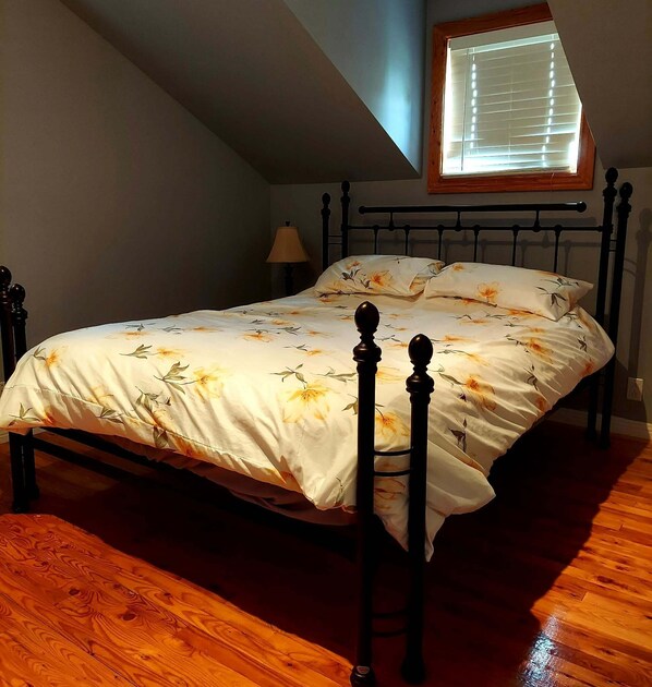 A bedroom with a bed, wooden floor, a window with blinds, and a lamp.