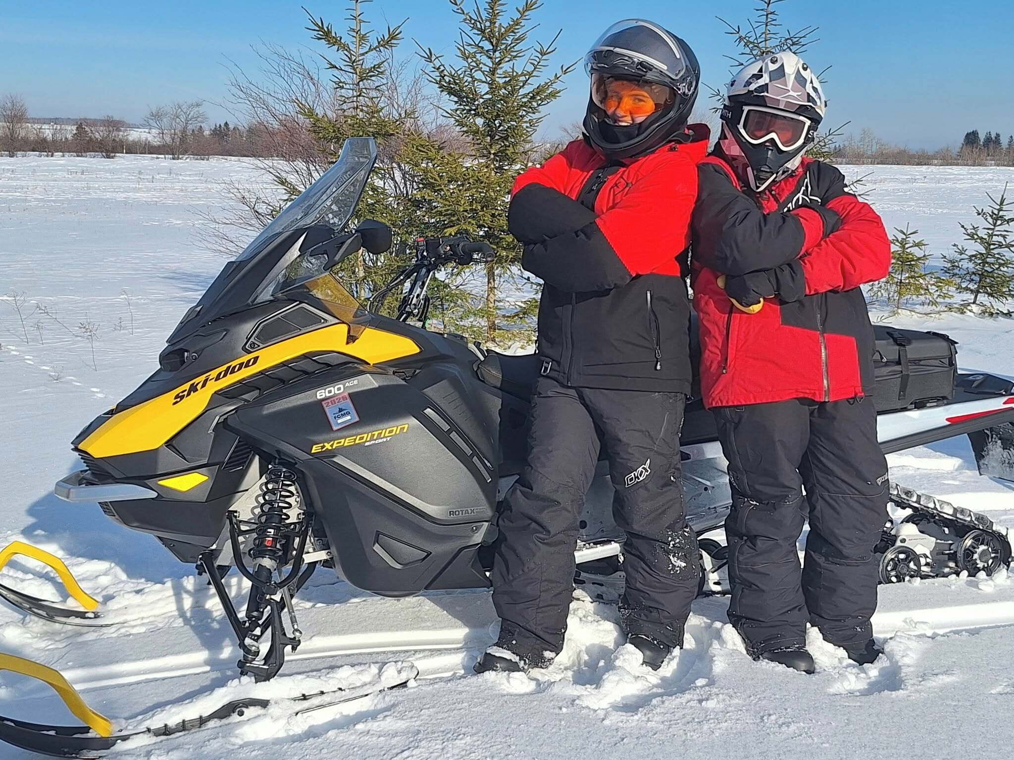 Dos personas de pie junto a un vehículo de nieve en un paisaje nevado.
