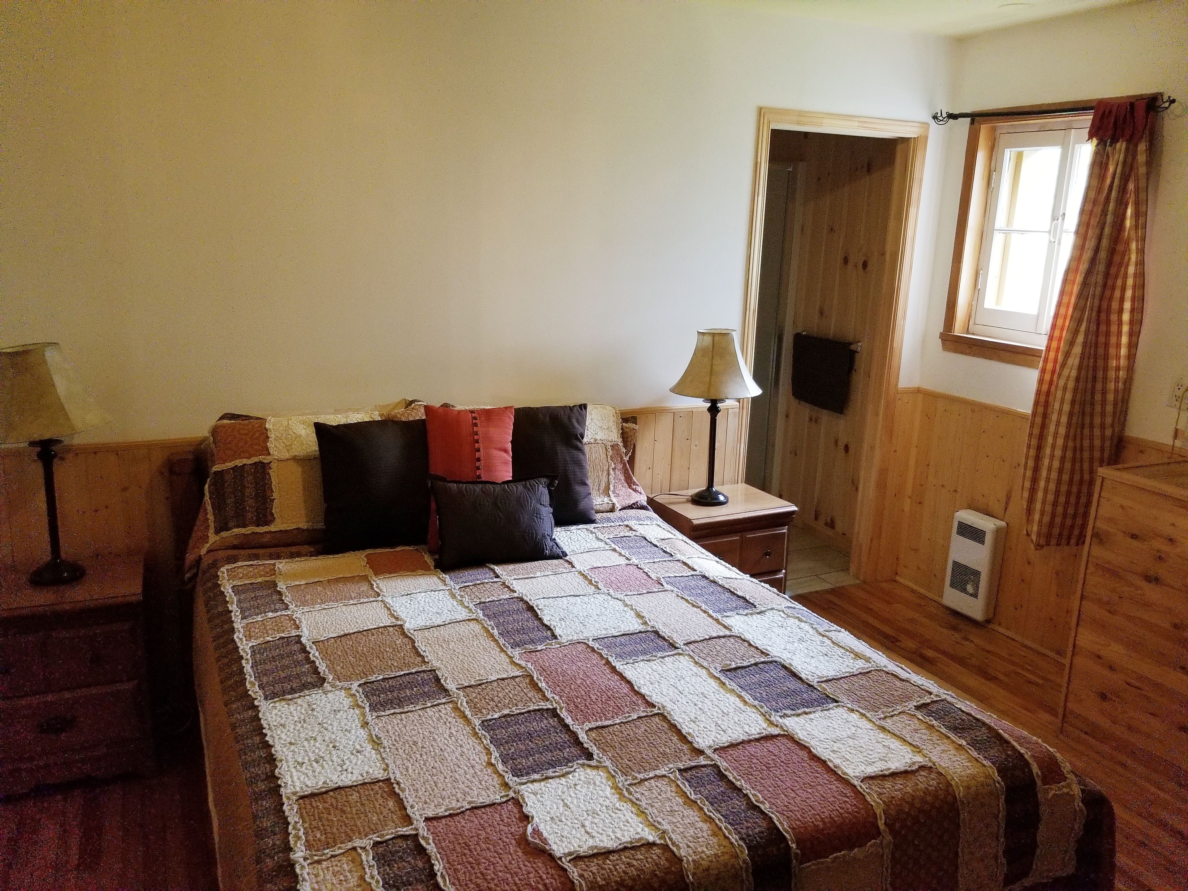 Quarto com piso de madeira, cama com colcha, mesa de cabeceira com abajur e janela com cortinas.