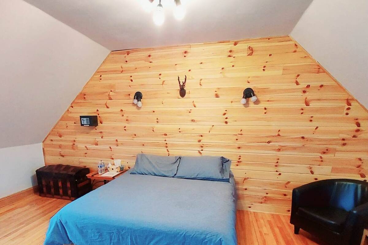Una camera da letto con soffitto spiovente, pannelli di legno a rivestimento, un letto con copriletto blu, una sedia nera e un tavolino con una lampada.