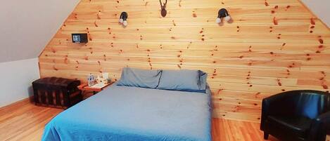 A bedroom with a sloped ceiling, wooden wall paneling, a bed with a blue bedspread, a black chair, and a small table with a lamp.