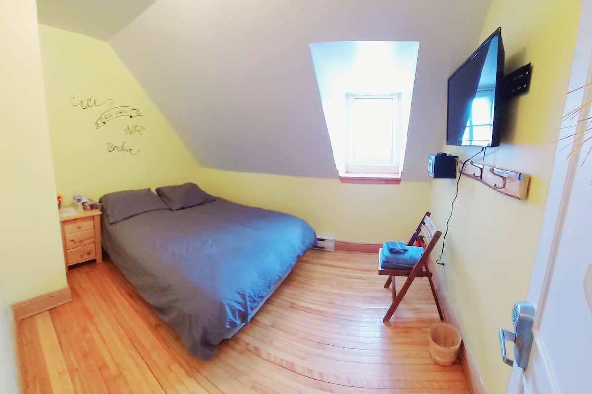 Una camera da letto accogliente e piccola con soffitto spiovente, un letto singolo, un tavolino, una sedia, un televisore montato a parete e un lucernario.