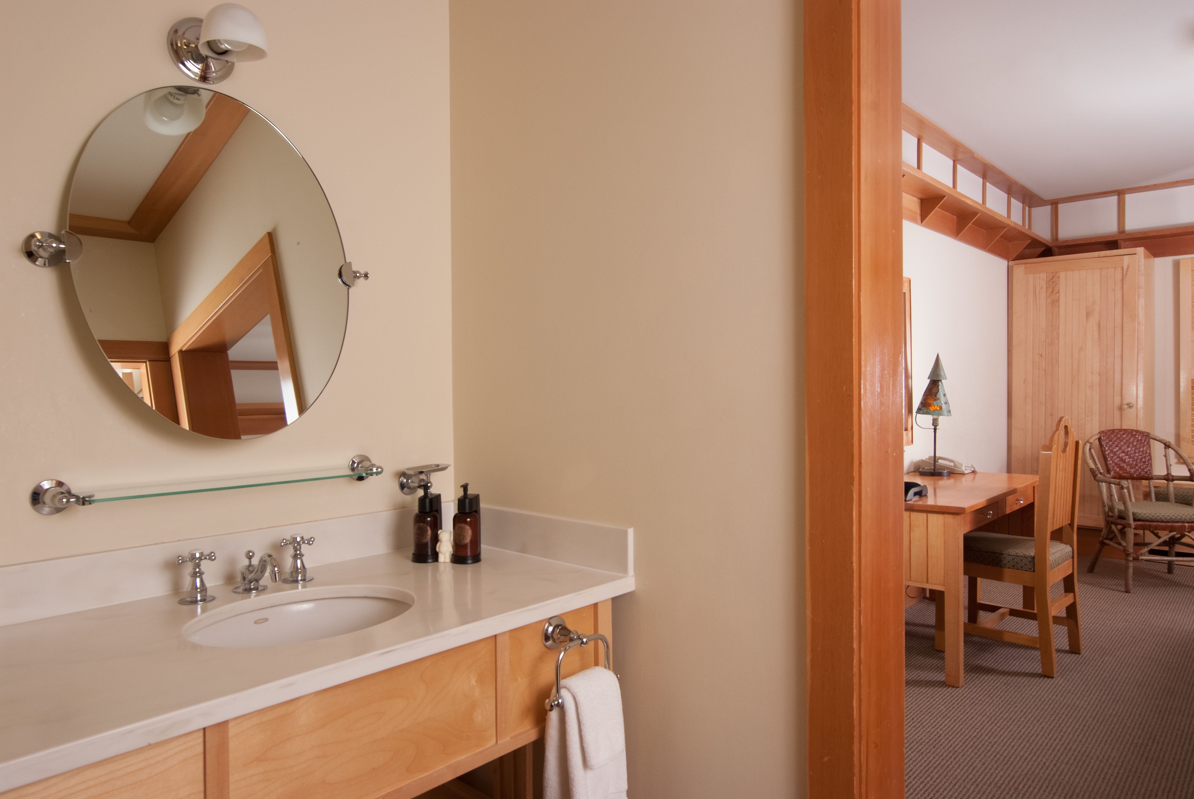 A bathroom with a round mirror, a sink, and a wooden vanity. There is a towel hanging on the wall and a chair in the adjacent room.