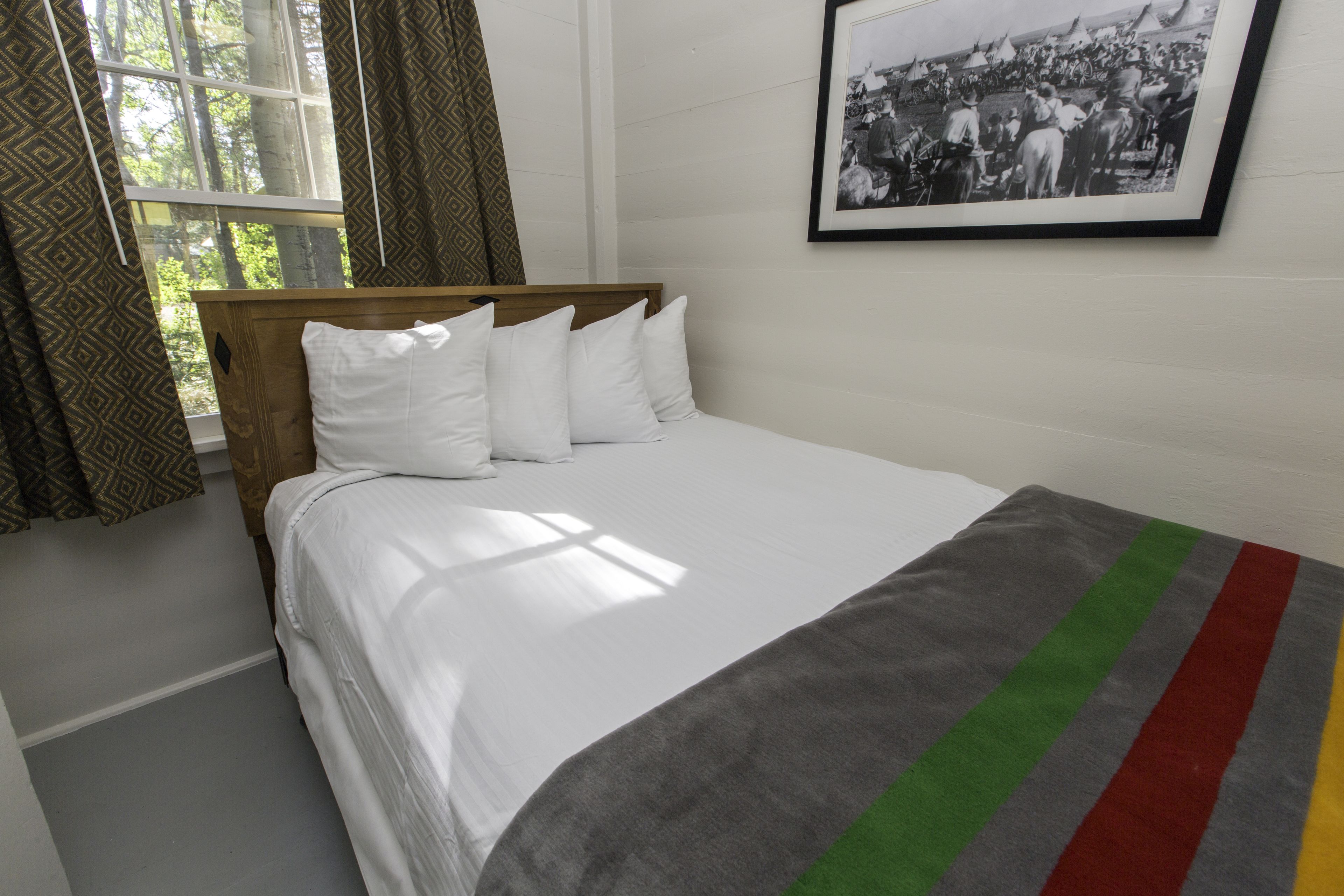 Ein ordentlich bezogenes Bett mit weißen Leinentüchern und einer gestreiften Decke, ein Fenster mit gemusterten Vorhängen und ein gerahmtes historisches Foto an der Wand.