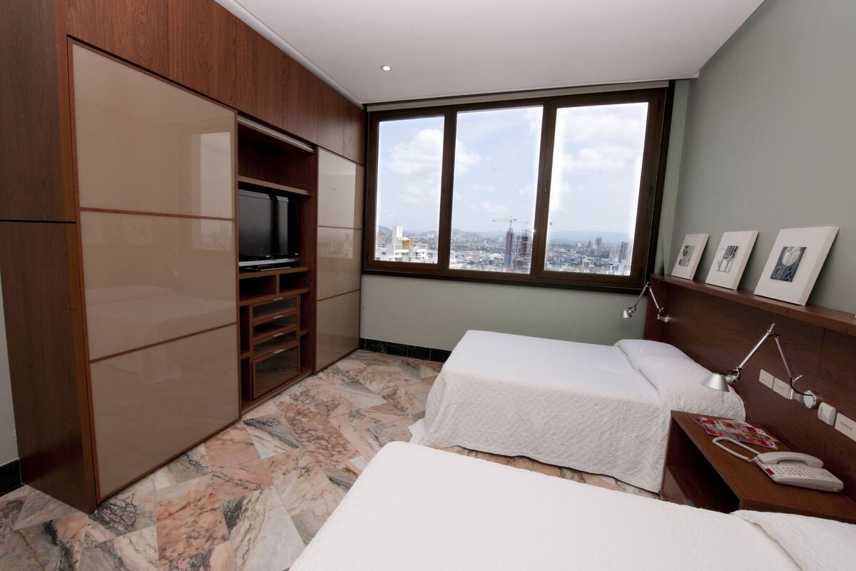 Ein Hotelzimmer mit zwei Betten, einem Fernseher, einem großen Fenster mit Blick auf die Stadt und einem Holzschrank.