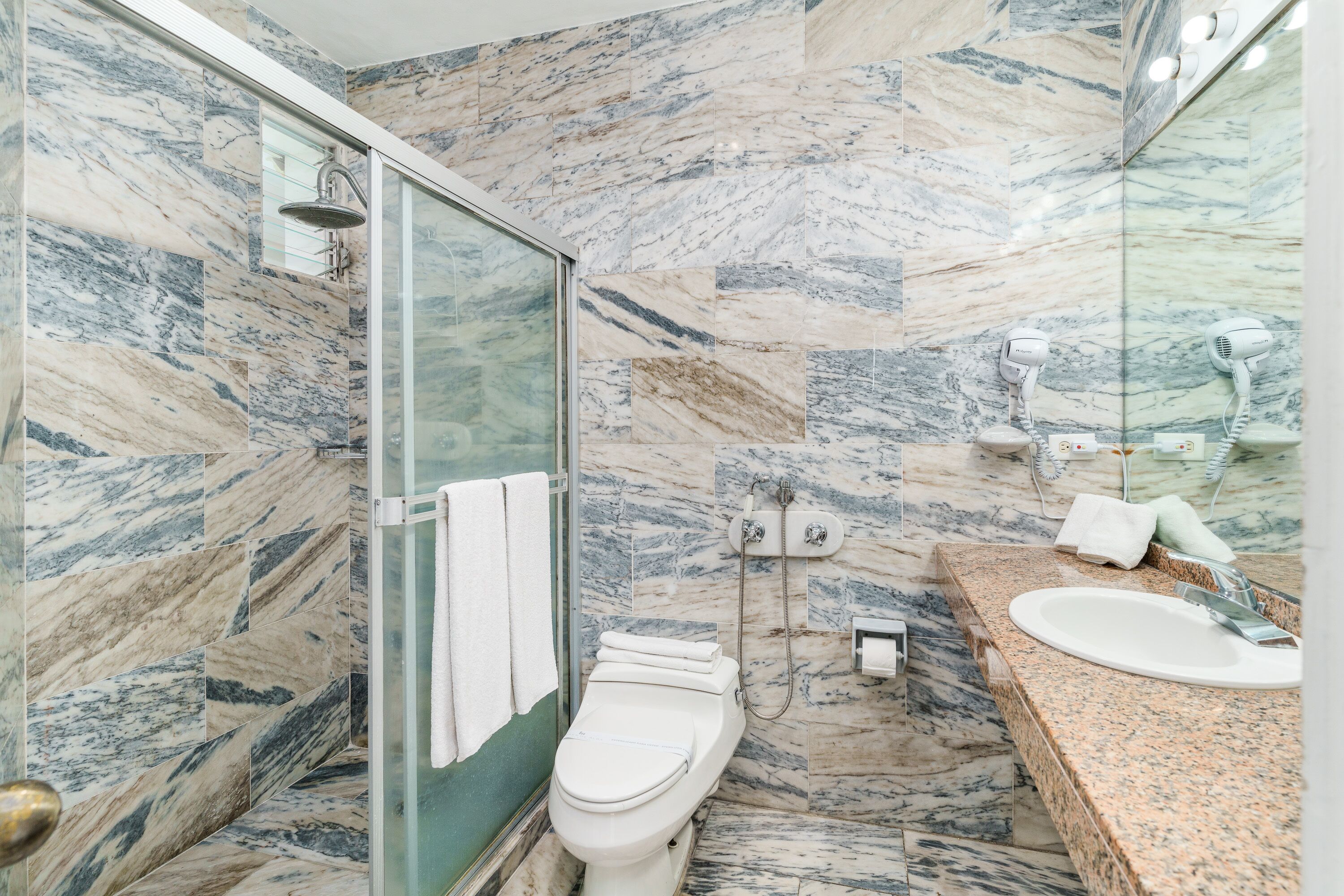 A bathroom with a glass shower enclosure, a toilet, a sink, and a marble-tiled wall.
