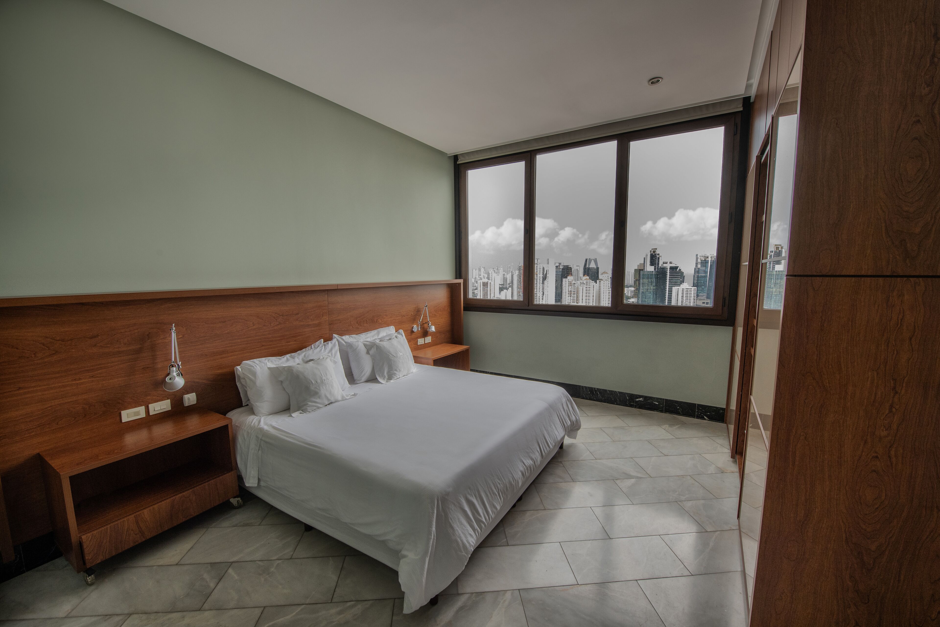 Une chambre d’hôtel moderne avec un grand lit, une tête de lit en bois, une table de chevet et une vue sur les toits de la ville, visible à travers une grande fenêtre.