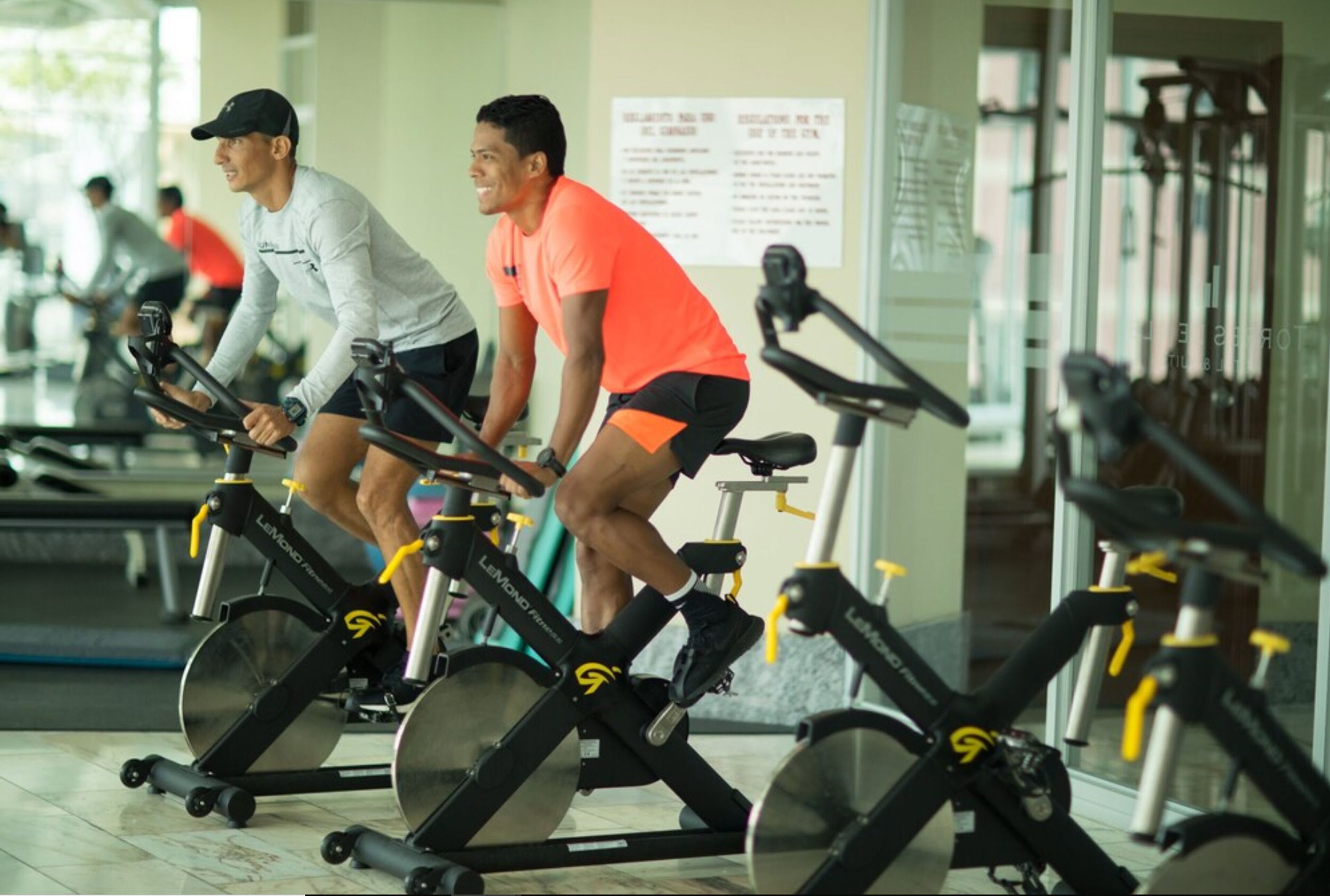 Deux personnes font du vélo stationnaire dans une salle de sport équipée de plusieurs vélos, et un panneau est accroché au mur.