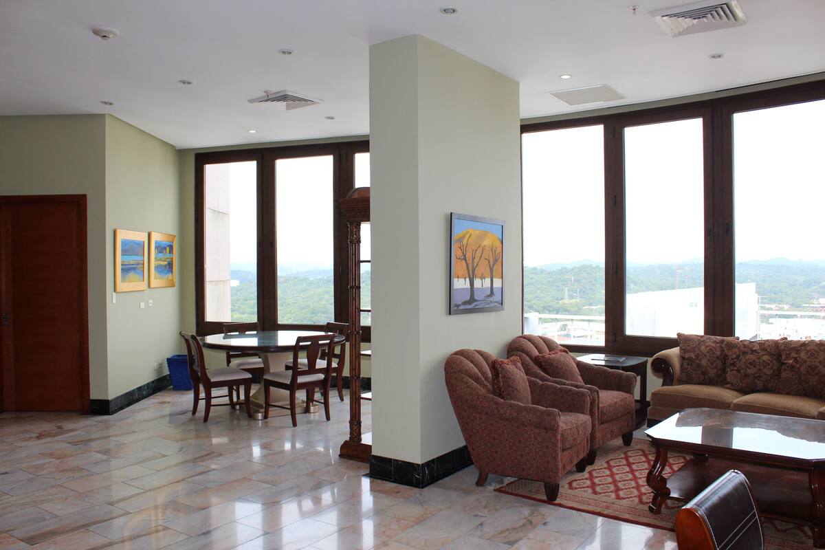 Ein geräumiger Wohnbereich mit Esstisch, mehreren Stühlen, einer Couch und großen Fenstern mit Blick auf die Landschaft.