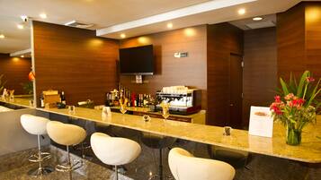 A bar area with a long counter, multiple stools, a television mounted on the wall, and a coffee machine.