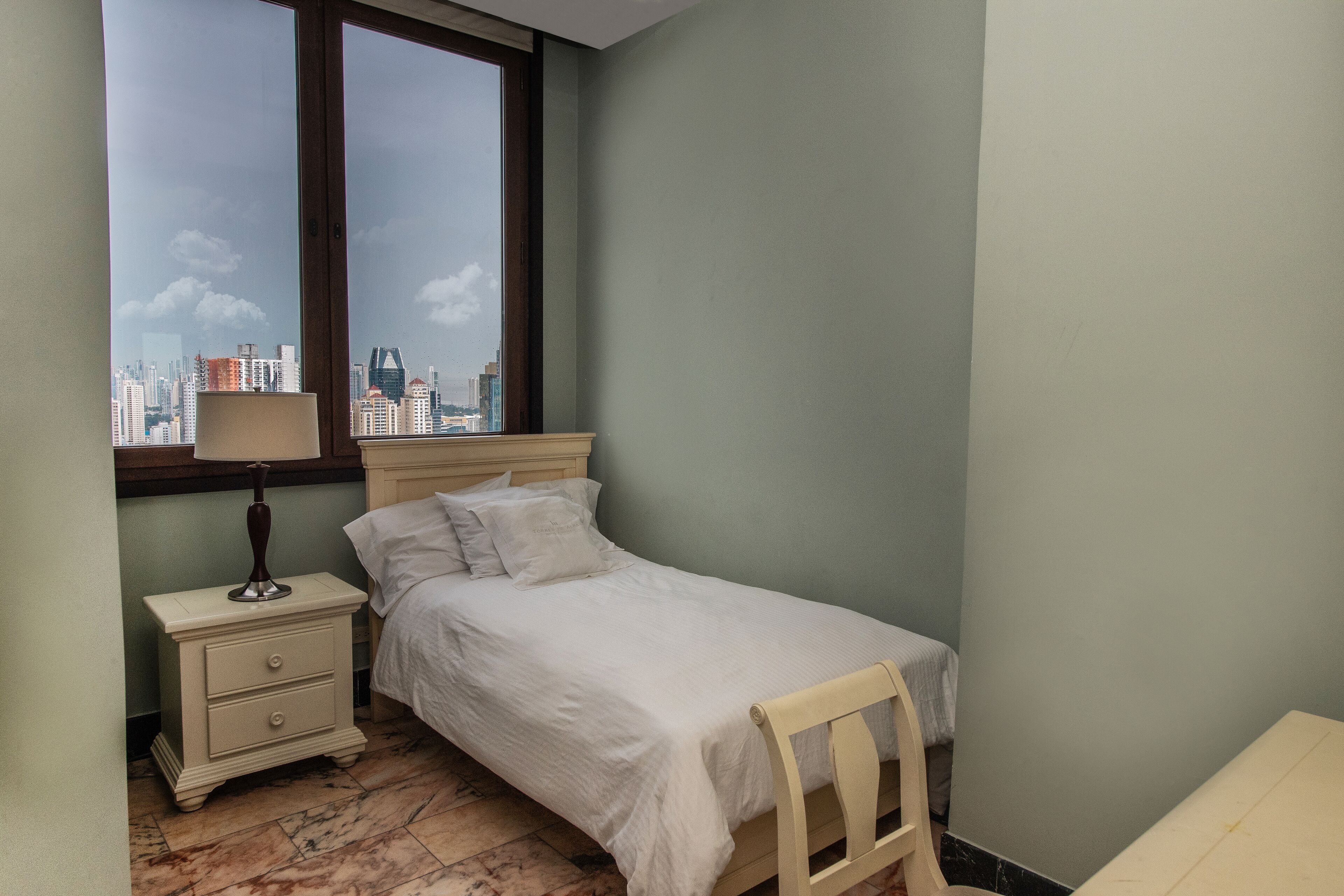 Ein Schlafzimmer mit Bett, Nachttisch, Lampe, Stuhl und Fenster mit Blick auf die Stadt.