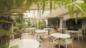 Un área de comedor al aire libre con mesas y sillas redondas, rodeada de plantas en macetas y una terraza de madera.