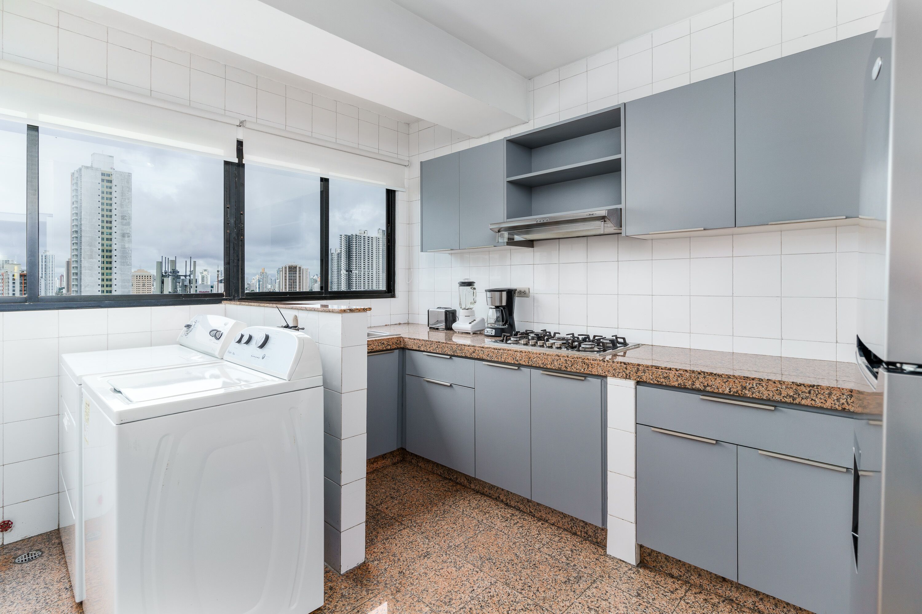 Una cocina moderna con lavadora y secadora, encimeras de granito, armarios grises y vistas a la ciudad a través del ventanal.