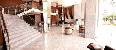 A spacious lobby with a marble floor, a staircase with glass railings, and a seating area with a coffee table and chairs.