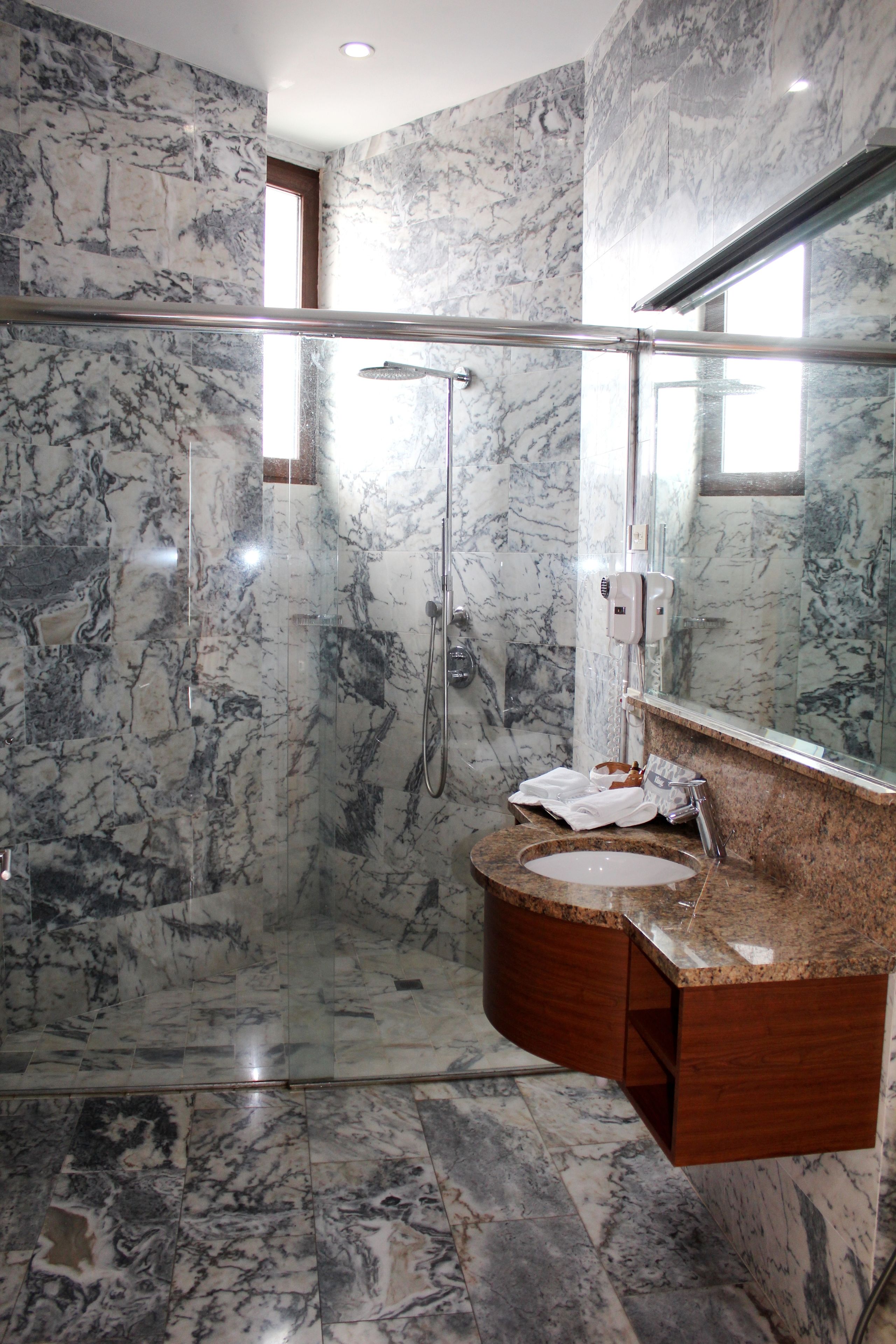Un baño con piso de mármol, ducha con mampara de vidrio, lavamanos con encimera de mármol y espejo sobre el lavamanos.