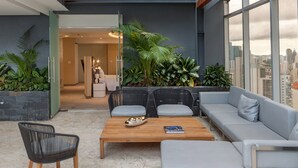 Un área de estar moderna con un sofá, sillas y una mesa de centro. Se ve la ciudad a través de amplias ventanas.