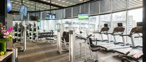 Un gimnasio moderno con máquinas de cardio, estaciones de pesas y grandes ventanales con vistas a la ciudad.