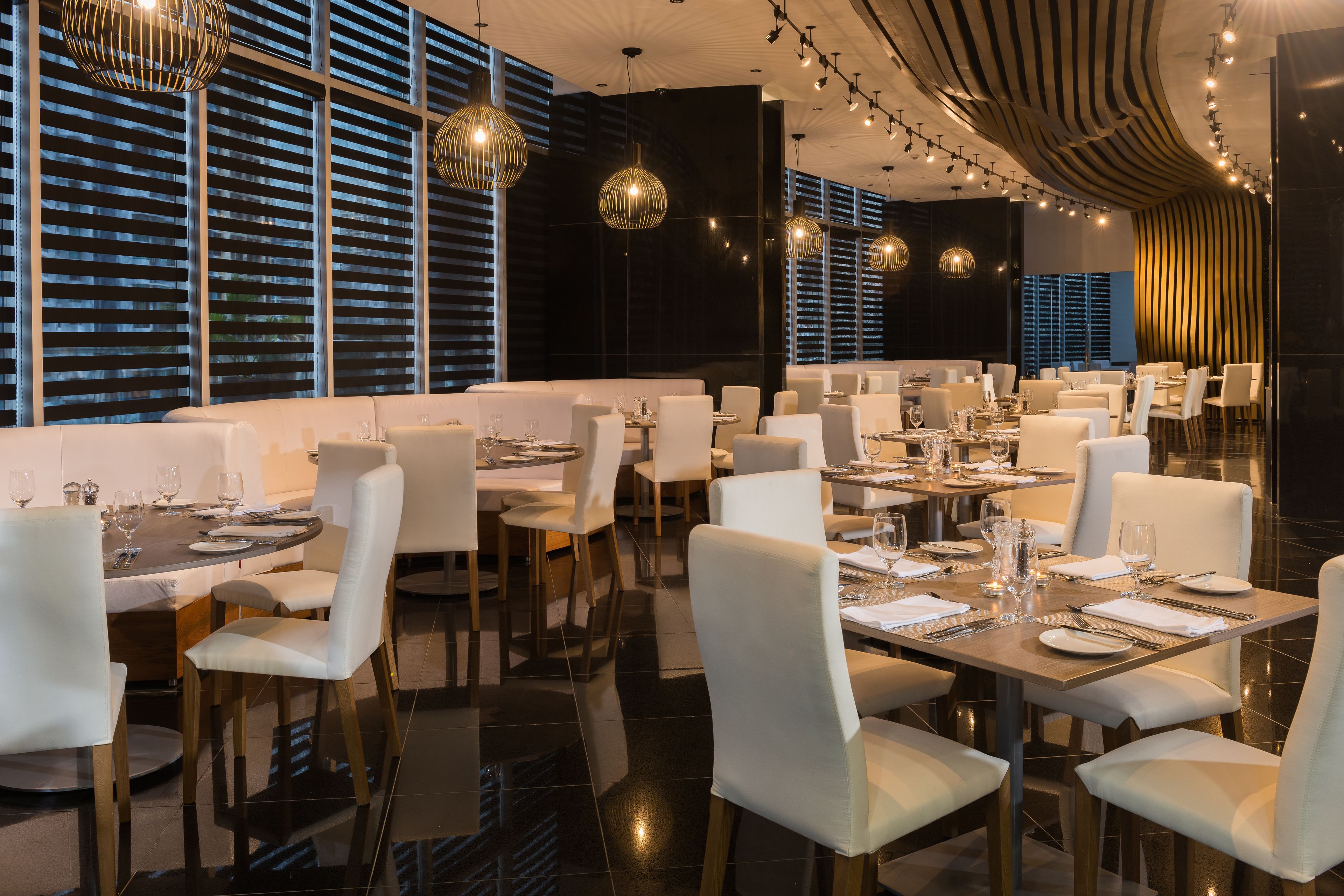 Un restaurant moderne avec des chaises et des tables blanches, de grandes fenêtres équipées de stores et un éclairage décoratif.