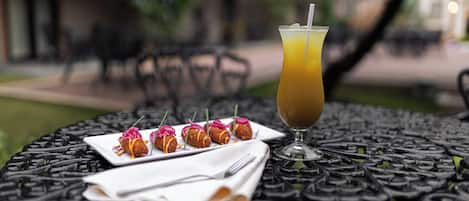 A table with a glass of orange juice, a plate of appetizers, and a napkin with cutlery on a black metal table.