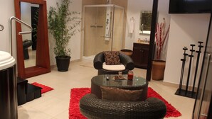 A modern bathroom with a glass shower enclosure, a black wicker chair, a glass coffee table, and a red rug. - Hotel Cubitá Boutique Resort & Spa (La Arena)