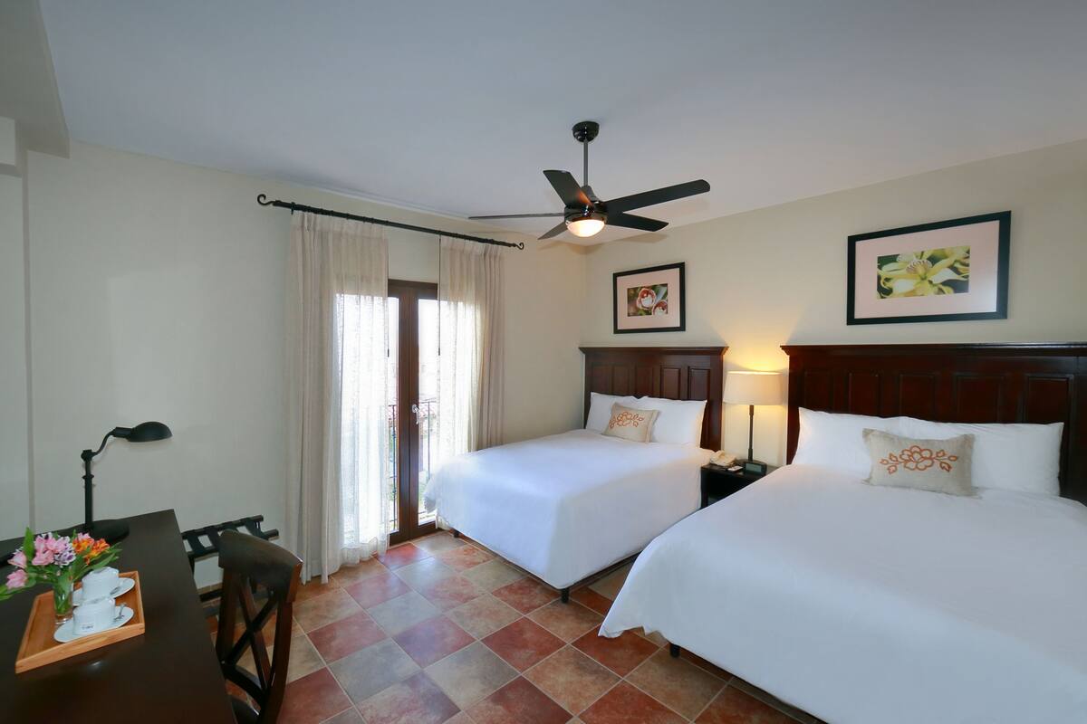 A hotel room with two beds, a ceiling fan, a desk with a lamp, and a window with curtains.