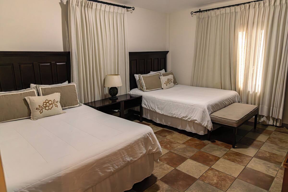 A hotel room with two beds, a nightstand, a lamp, and a window with curtains.