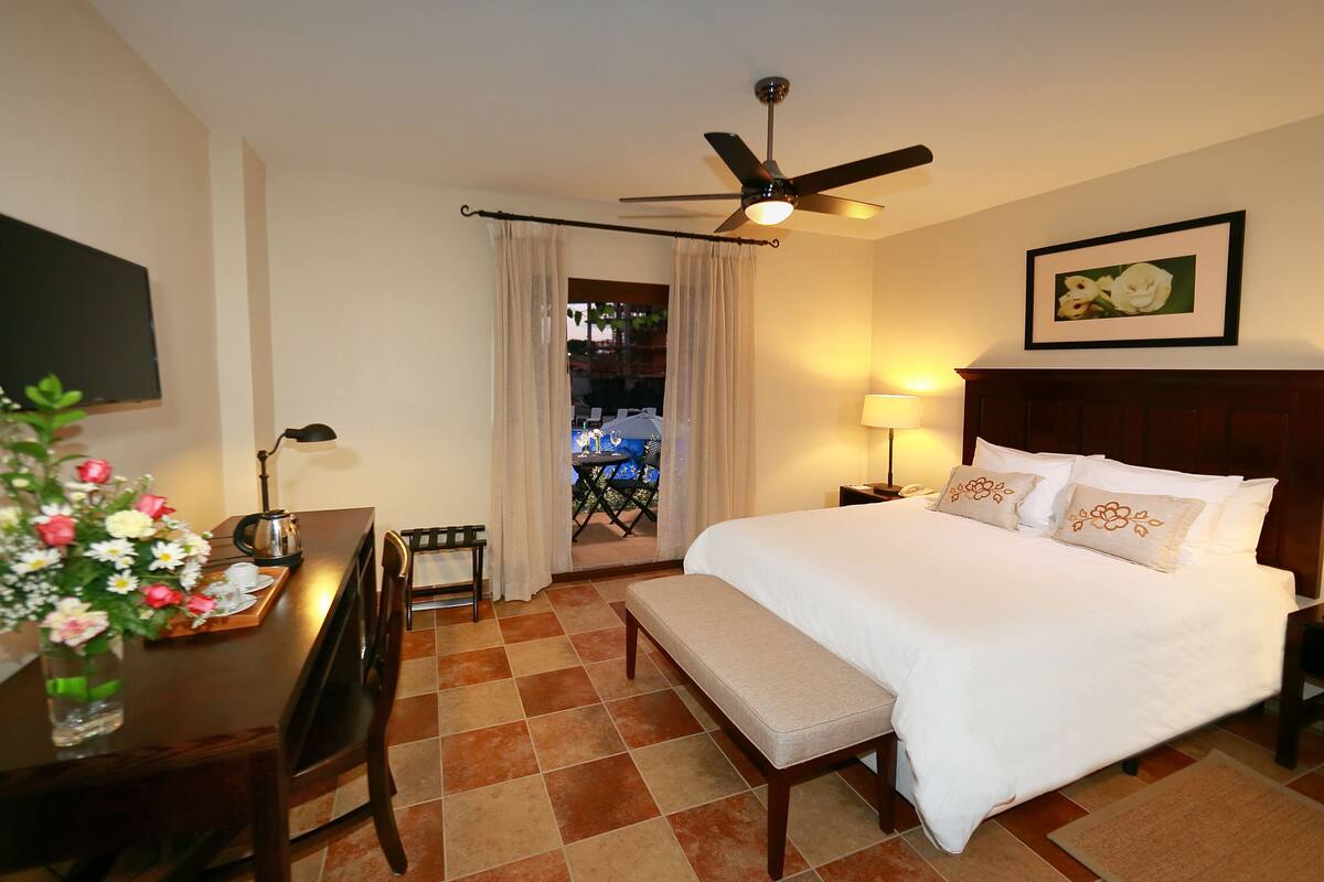 A hotel room with a bed, a desk with a lamp, a ceiling fan, and a view of the outdoors.