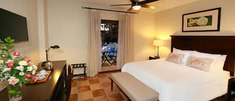 A hotel room with a bed, a desk with a lamp, a ceiling fan, and a view of the outdoors.
