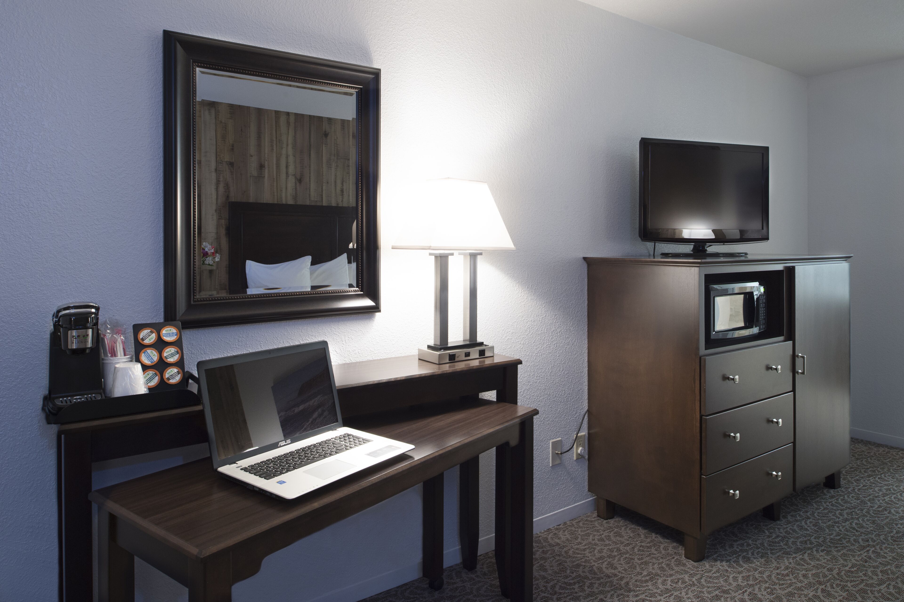 A hotel room with a wooden desk, a laptop, a television, a lamp, and a minibar.