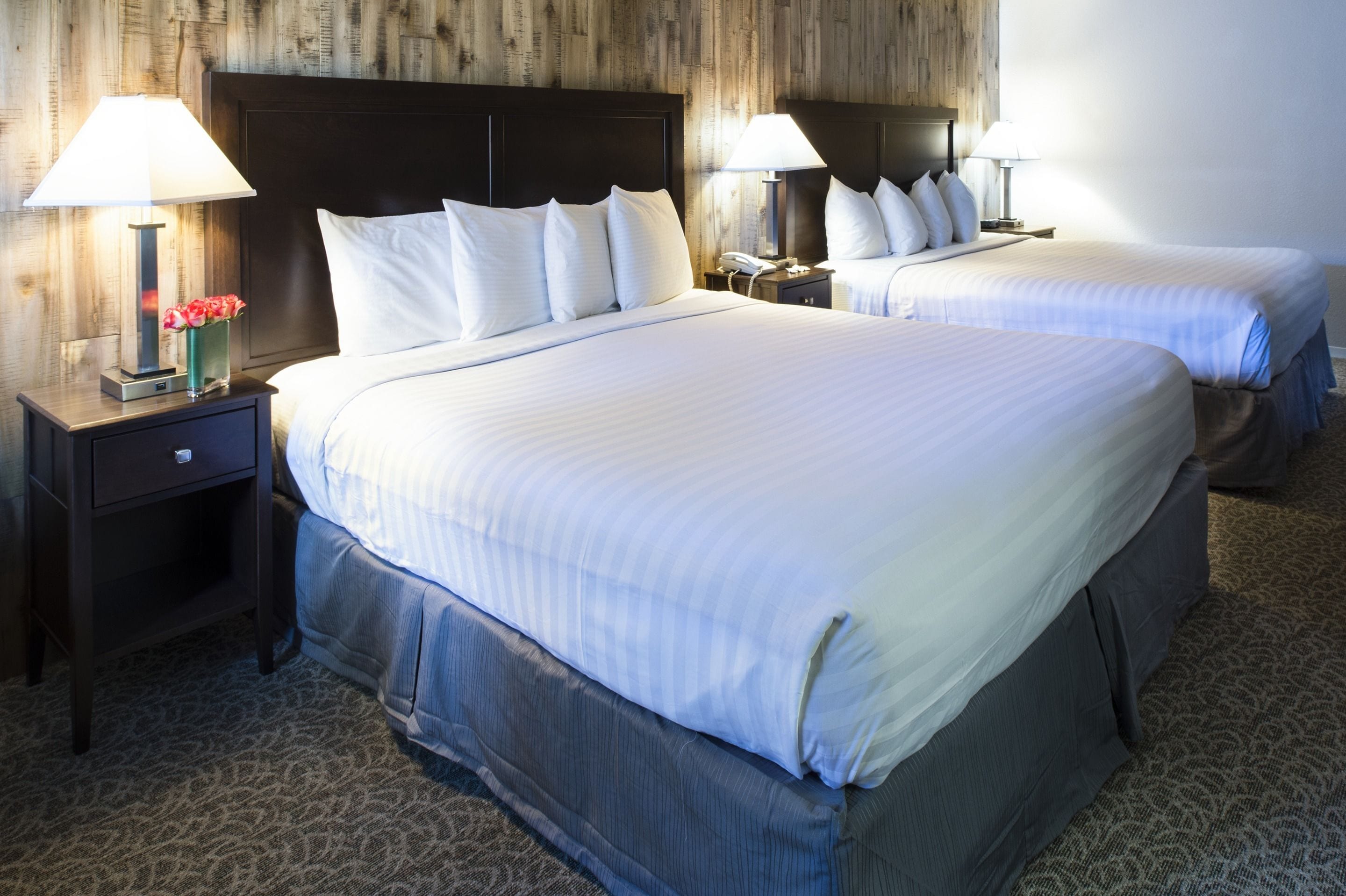 Una habitación de hotel con dos camas, cada una con ropa blanca y cabeceros oscuros. Hay mesitas de noche con lámparas y un florero con flores.