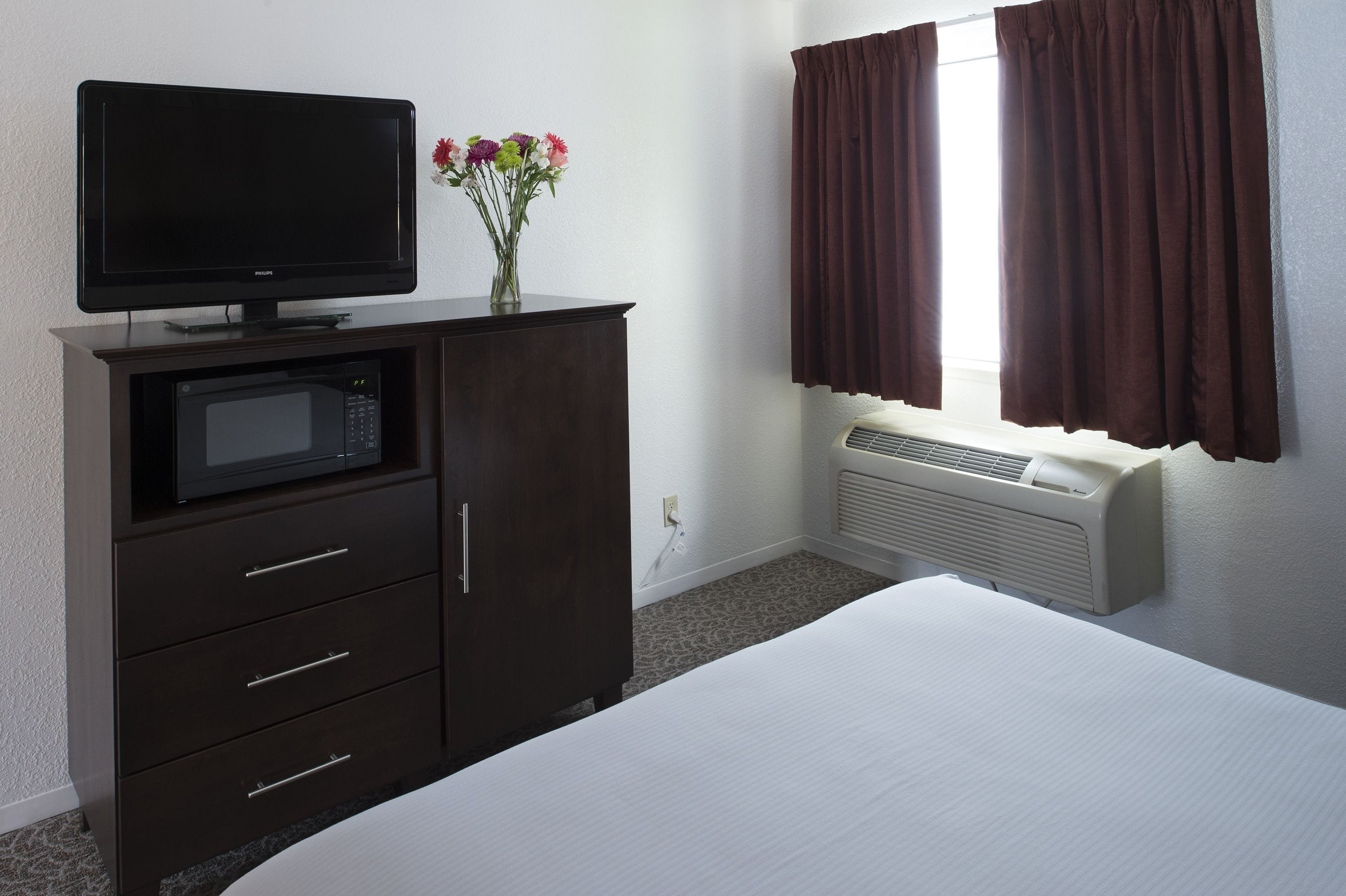 Una habitación de hotel con una cama, un mueble de madera oscura con televisor de pantalla plana y microondas, un jarrón con flores y una ventana con cortinas marrones.