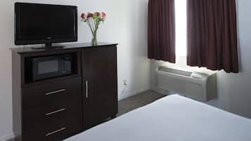 Una habitación de hotel con una cama, un mueble de madera oscura con televisor de pantalla plana y microondas, un jarrón con flores y una ventana con cortinas marrones.